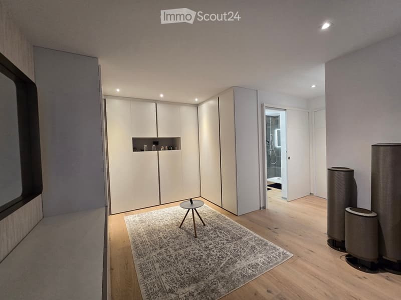 Modern, minimalist room with light wooden flooring, a patterned rug, a small round stool, built-in white wardrobes, and two tall speakers on the floor.