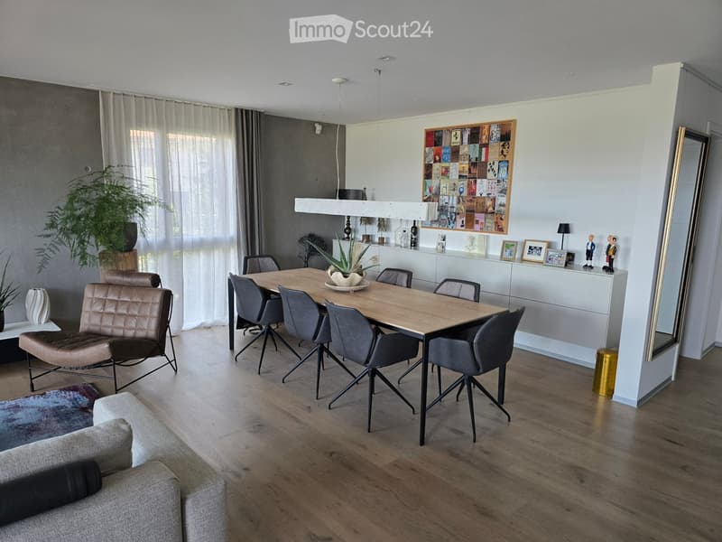 Modern living and dining area with a grey sofa, leather armchair, wooden dining table, grey chairs, large mirror, and decorative plants.