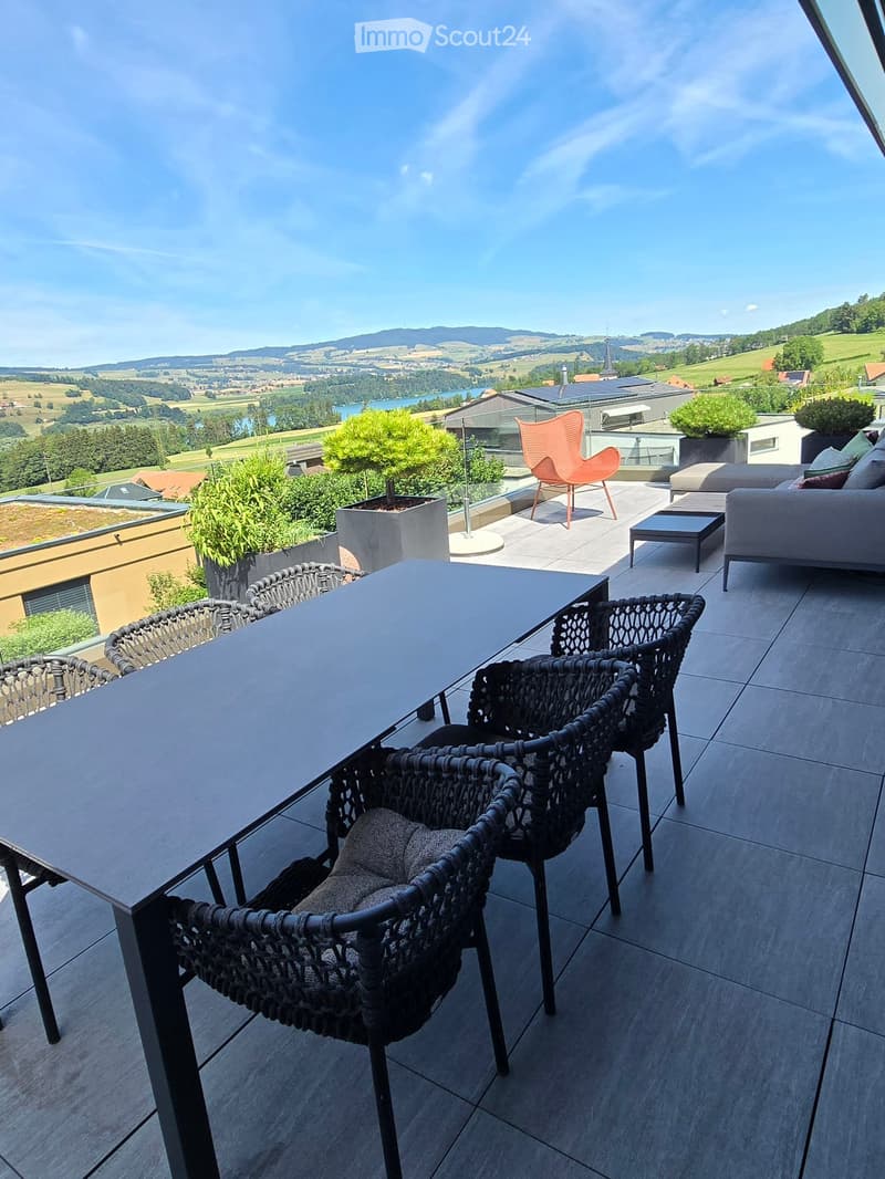Balcony with a scenic view, featuring a table with chairs, a sofa, and a stool, surrounded by potted plants.