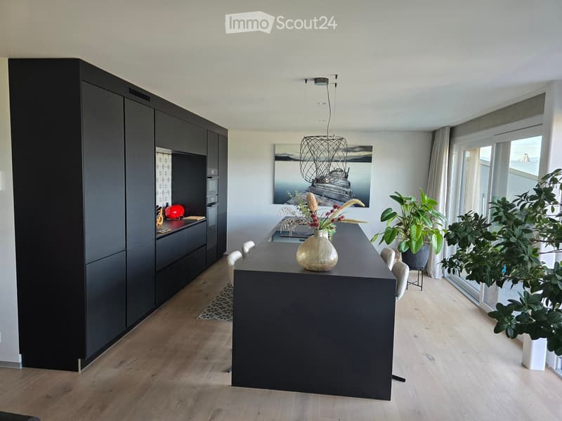Modern kitchen with dark cabinets, a long black counter, and a vase with dried flowers on the counter. There are chairs around the counter, a large window with a view, and several potted plants near the window.