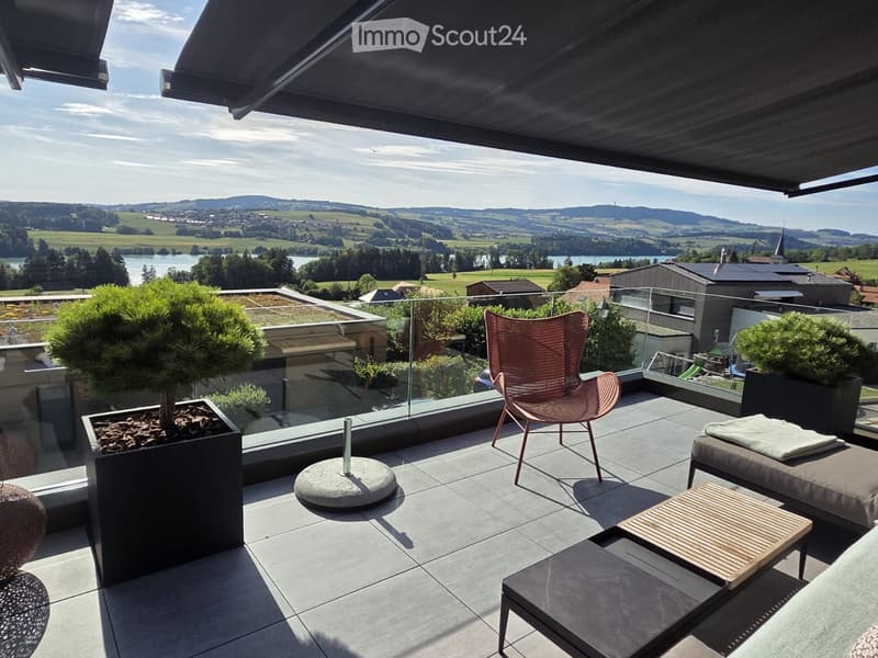 Modern balcony with glass railing, outdoor furniture including a chair, table, and potted plants, overlooking a scenic landscape with a lake and hills.