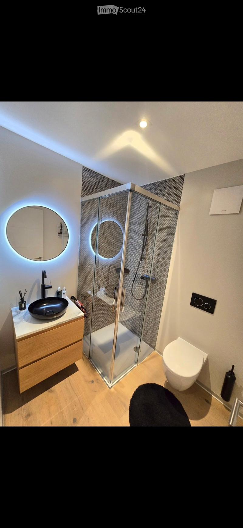 Modern bathroom with a wooden vanity, black sink, round illuminated mirror, glass-enclosed shower, and toilet.