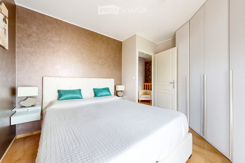Modern bedroom with a large bed, white headboard, teal pillows, nightstands with lamps, and an open door leading to another room with a chair and small table.