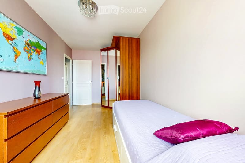 Bright bedroom with wooden flooring, a neatly made bed with a single pillow, a wooden dresser with a decorative vase, a large mirror, and a wooden wardrobe. A world map is mounted on the wall.
