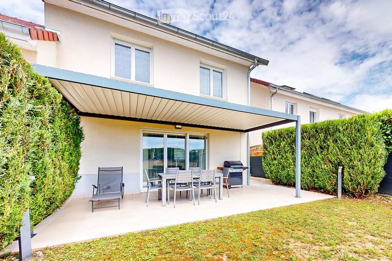 Outdoor patio area with a covered seating area, a dining table with chairs, and a grill, surrounded by neatly trimmed hedges and a lawn.