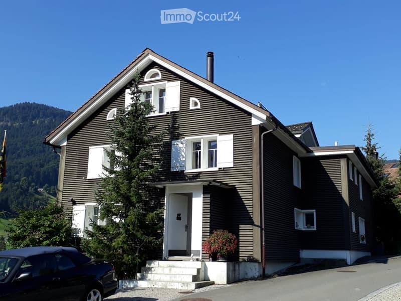 Two-story house with dark brown siding, white shutters, and a raised ground floor entrance. A small garden area with potted plants is visible near the entrance.