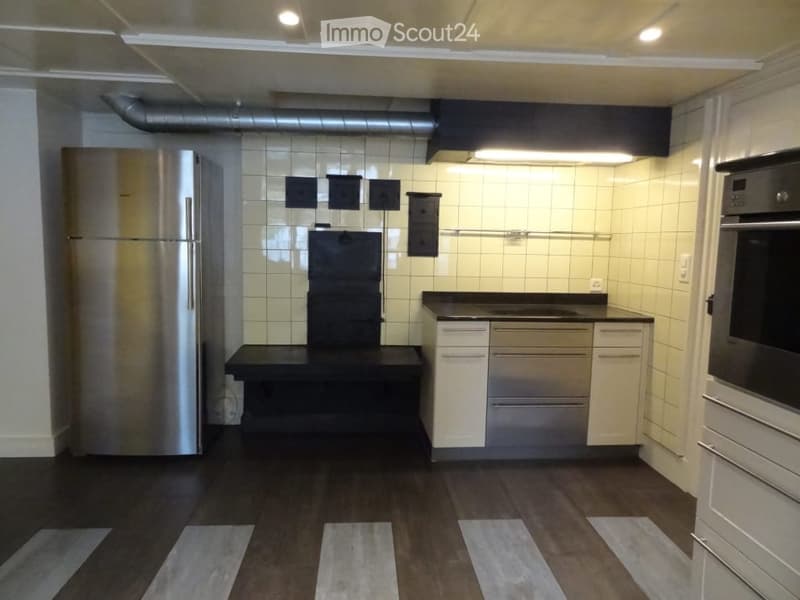 Modern kitchen with stainless steel appliances, white tiled walls, and dark wooden flooring. Features include a refrigerator, oven, and built-in cabinets.