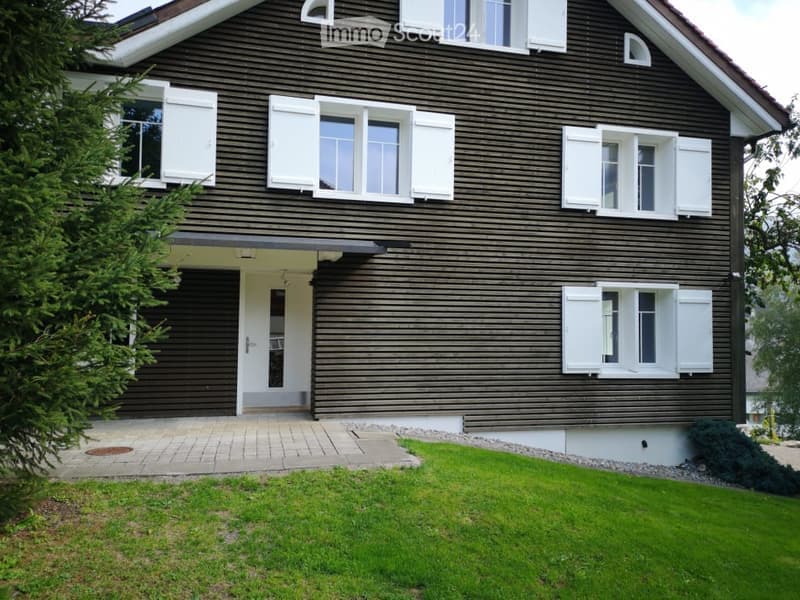 Two-story house with dark brown wooden siding, white shutters, and multiple windows. Features a covered entrance with a small porch and a well-maintained lawn.