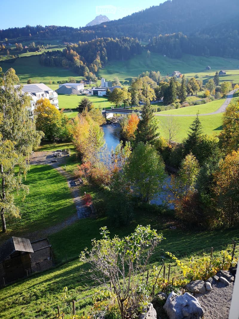 Scenic view of a lush green valley with a river, scattered houses, and rolling hills under a clear blue sky.