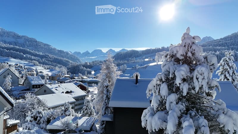 Snow-covered village with houses, trees, and mountains in the background under a clear blue sky.
