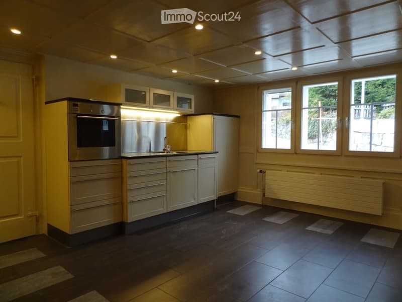 Modern kitchen with stainless steel oven, light wood cabinets, and dark tiled floor. Three windows provide natural light.