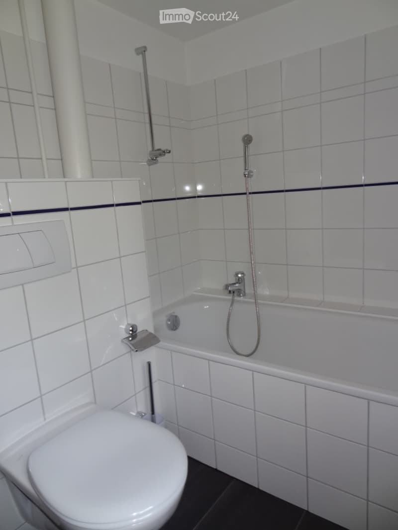 Modern bathroom with white tiled walls, a toilet, a bathtub with a handheld shower hose, and a built-in shelf above the bathtub.