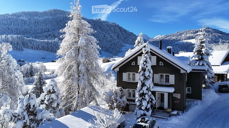 A two-story wooden house with white shutters and a snow-covered roof, surrounded by snow-laden trees and a snowy landscape.