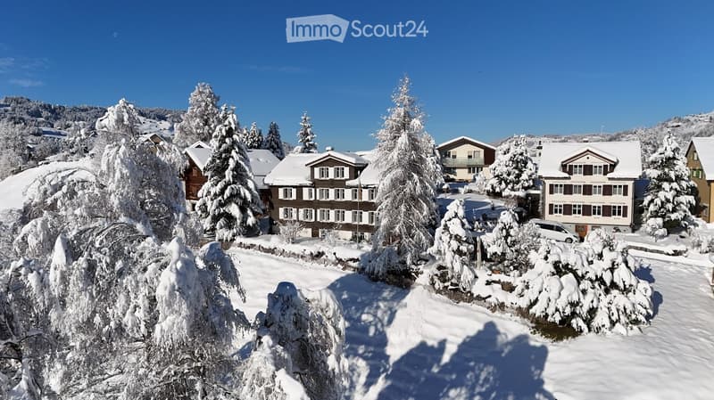 A snow-covered village with traditional houses, surrounded by snow-laden trees and mountains under a clear blue sky.