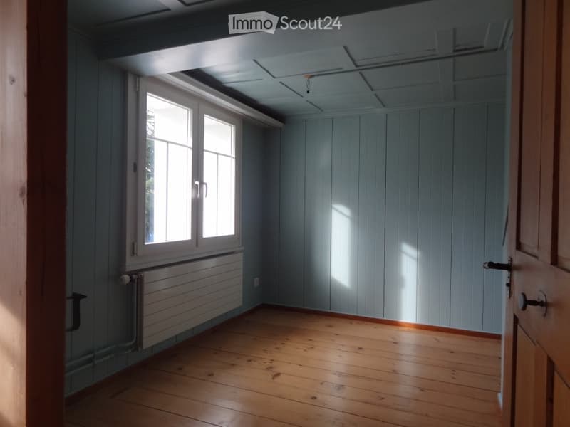 Empty room with wooden flooring, light blue paneled walls, and large windows allowing natural light to enter. The room features a radiator under the windows and a door on the right side.