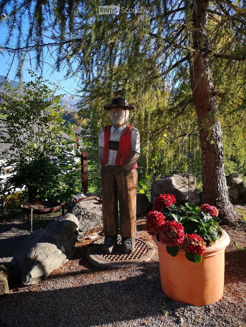 Garden area with a wooden statue of a man in work attire, a large terracotta flower pot with red flowers, and surrounding rocks and trees.