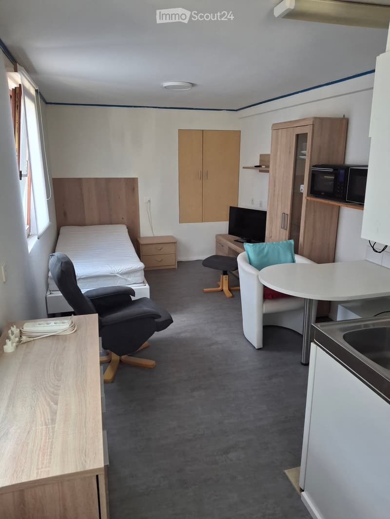 A compact, furnished room with a bed, desk, chair, microwave, and small kitchen area.