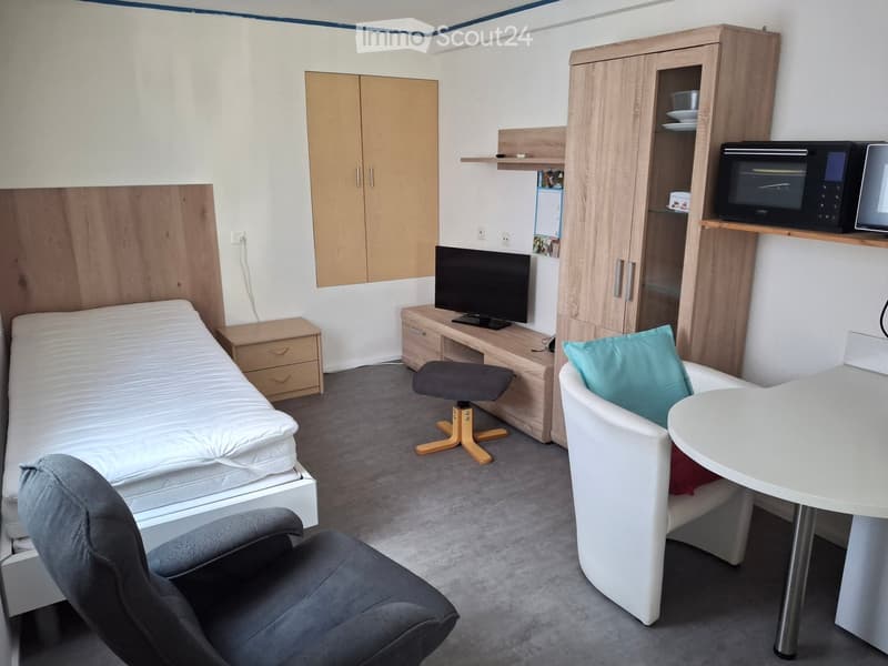 A compact, furnished room with a single bed, a small table and chair, a microwave, a cabinet, and a TV stand with a screen.
