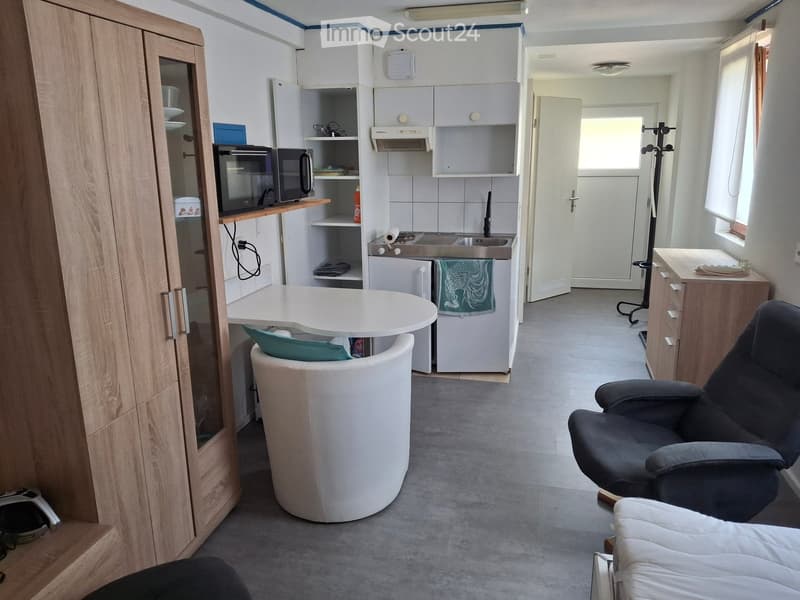 A compact, well-furnished room combining a living and kitchen area. Features a sofa, chair, small round table, microwave, refrigerator, kitchen cabinets, sink, and a door leading to another room.