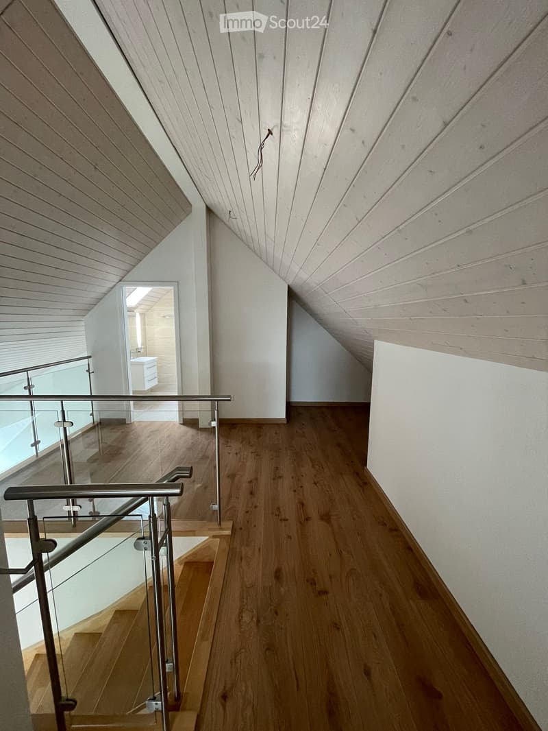 Modern hallway with wooden flooring, white walls, and a sleek metal railing leading to a staircase. The ceiling features a geometric wooden design.