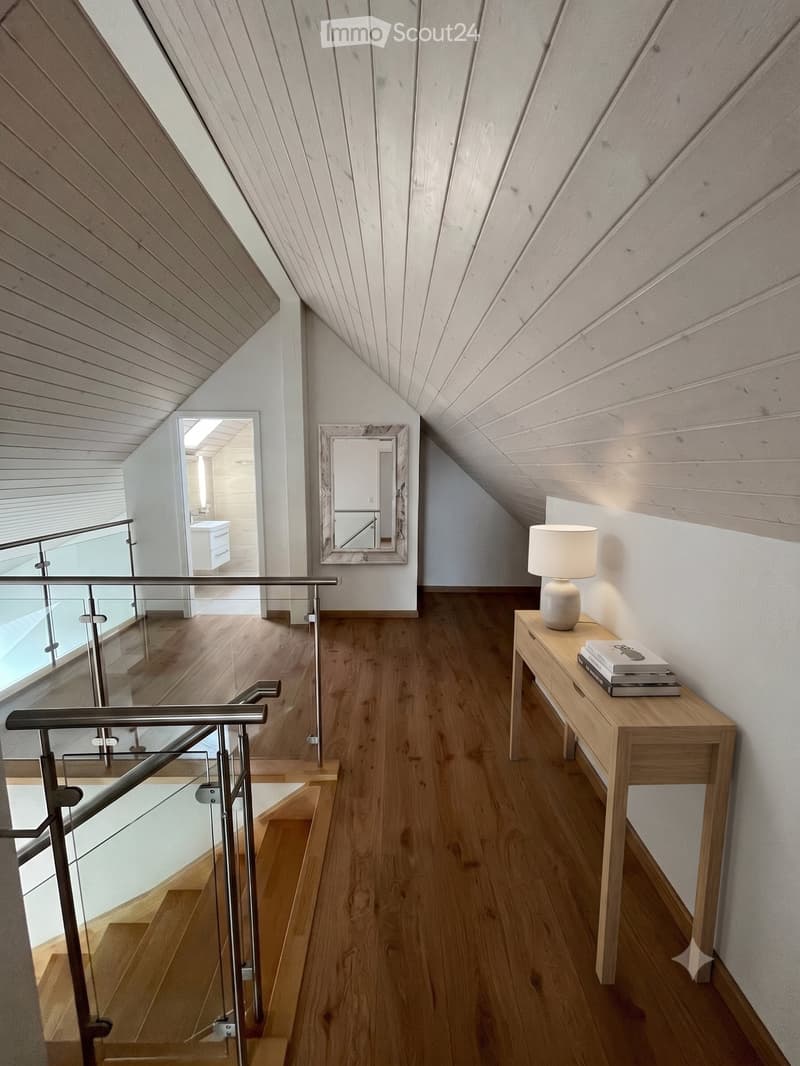 Modern hallway with wooden flooring, slanted wooden ceiling, glass railing, small table with lamp and books, and a mirror on the wall.