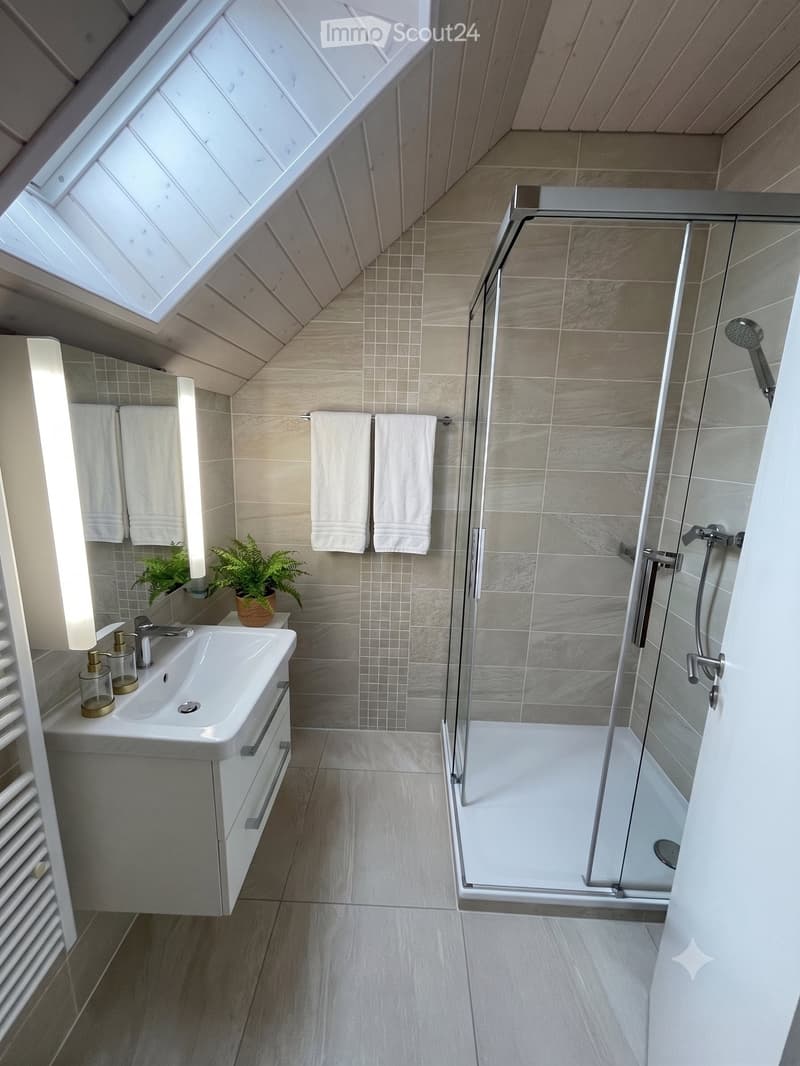 Modern bathroom with a white sink, glass-enclosed shower, and two white towels hanging on the wall. The room features light-colored tiles and a skylight window.
