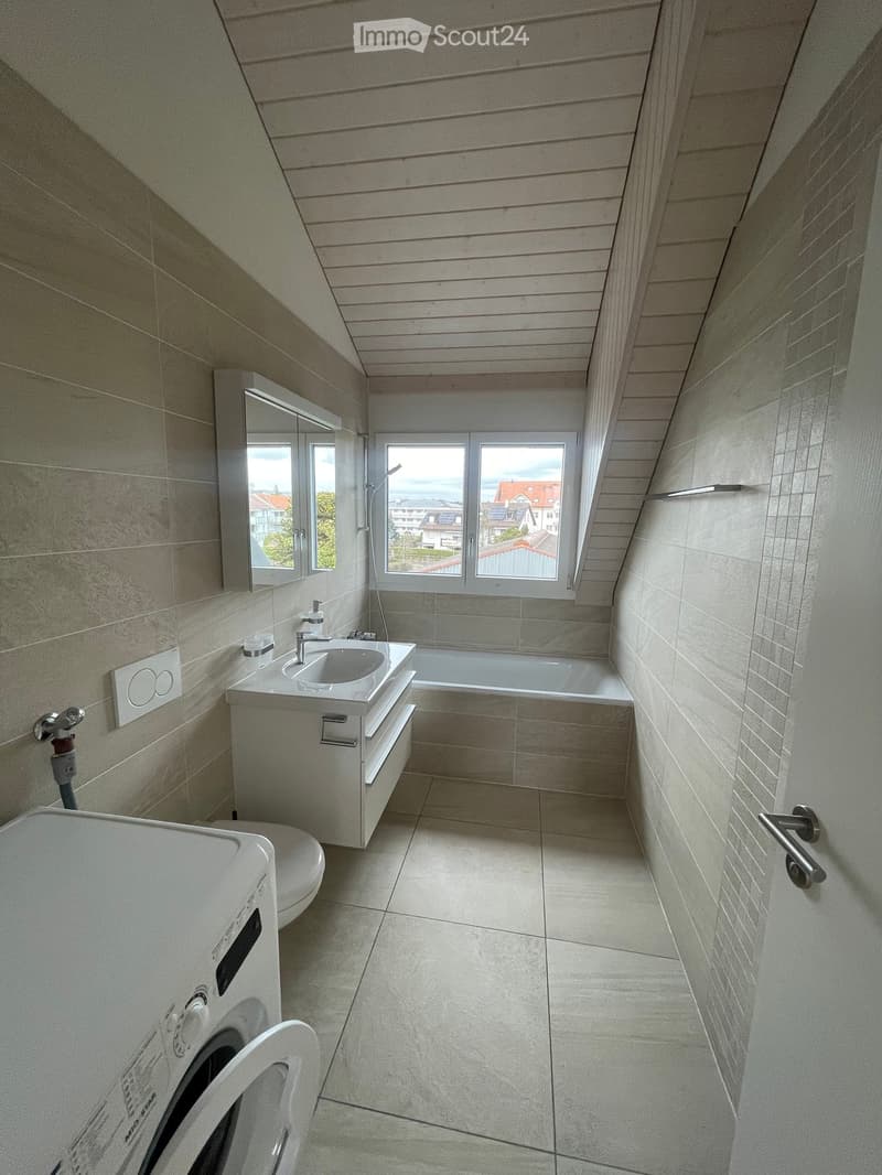 Modern bathroom with a sink, bathtub, toilet, and mirror. The room features light-colored tiles and a slanted wooden ceiling.