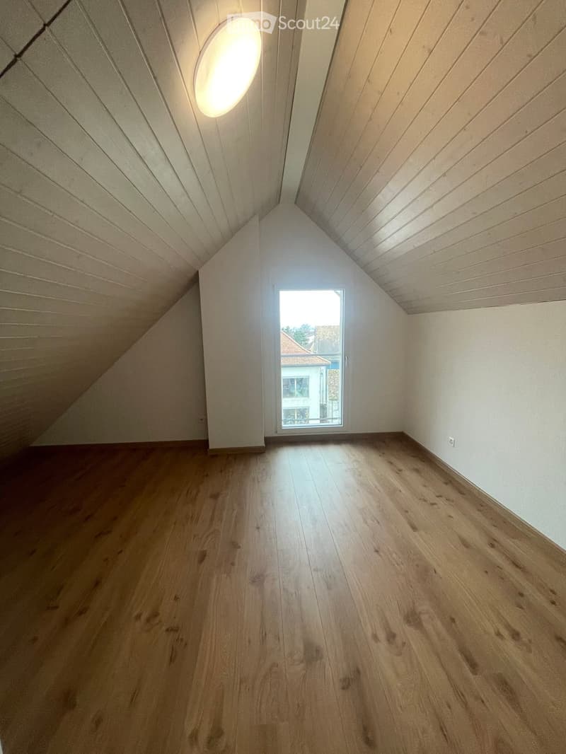 Empty attic room with wooden flooring, white walls, and a skylight window offering a view of the outside.