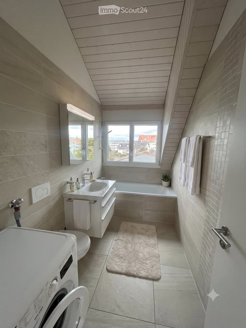 Modern bathroom with a sink, bathtub, toilet, mirror, and a small plant on the windowsill. The room features light-colored tiles and a slanted wooden ceiling.