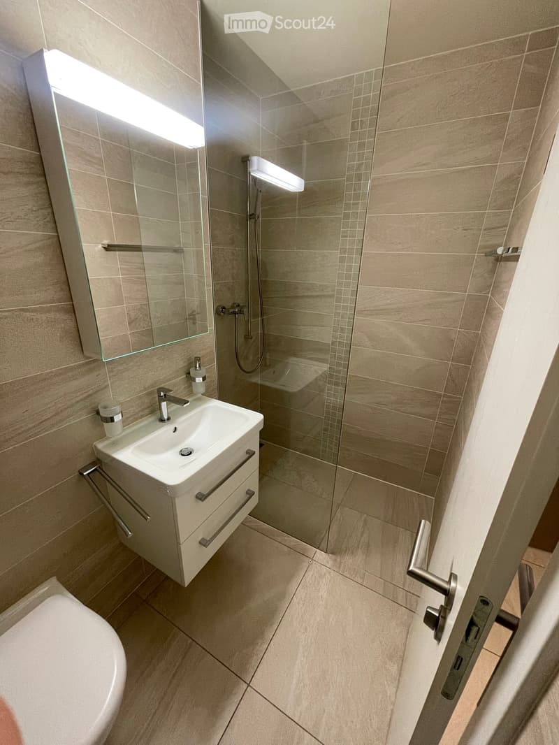 Modern bathroom with beige tiles, a white sink with a chrome faucet, a large mirror, a glass-enclosed shower, and a toilet.
