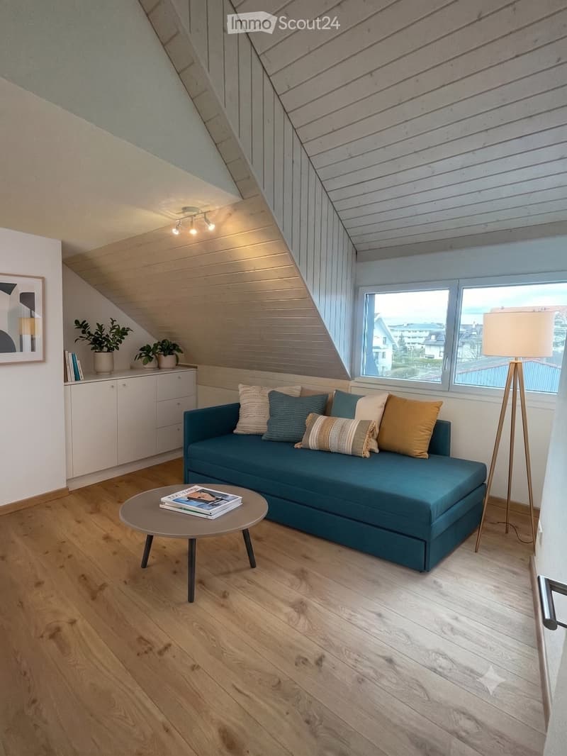 Modern living room with a teal sofa, wooden floor, large window, and decorative items including a floor lamp, coffee table, and potted plants.