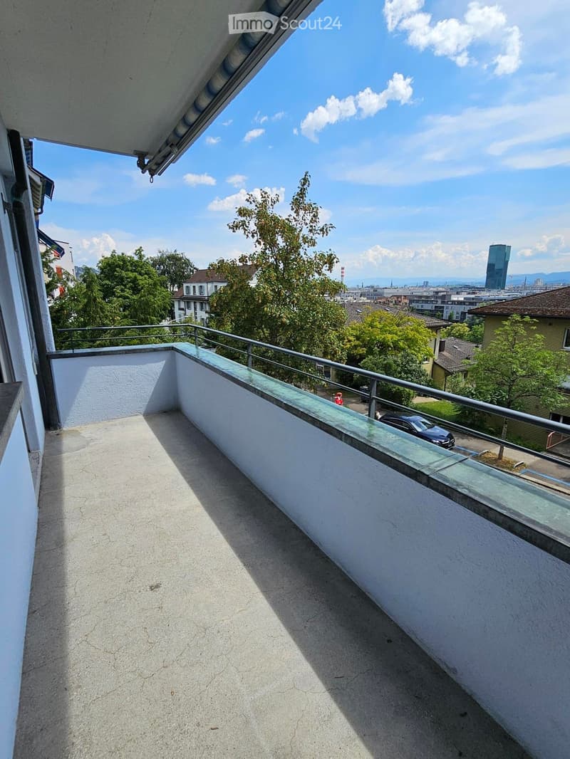Balcony with a modern glass railing, overlooking a cityscape with trees and buildings.