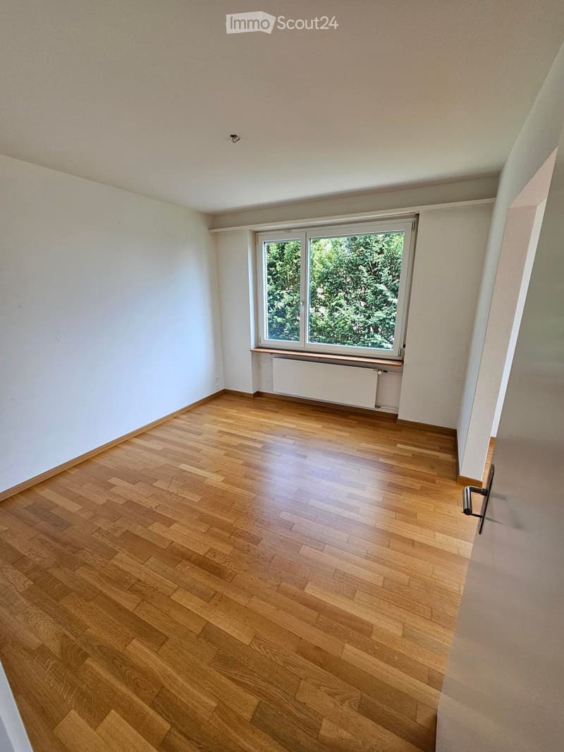 Empty room with wooden flooring, white walls, and a large window with a view of greenery. No furniture or equipment is present.