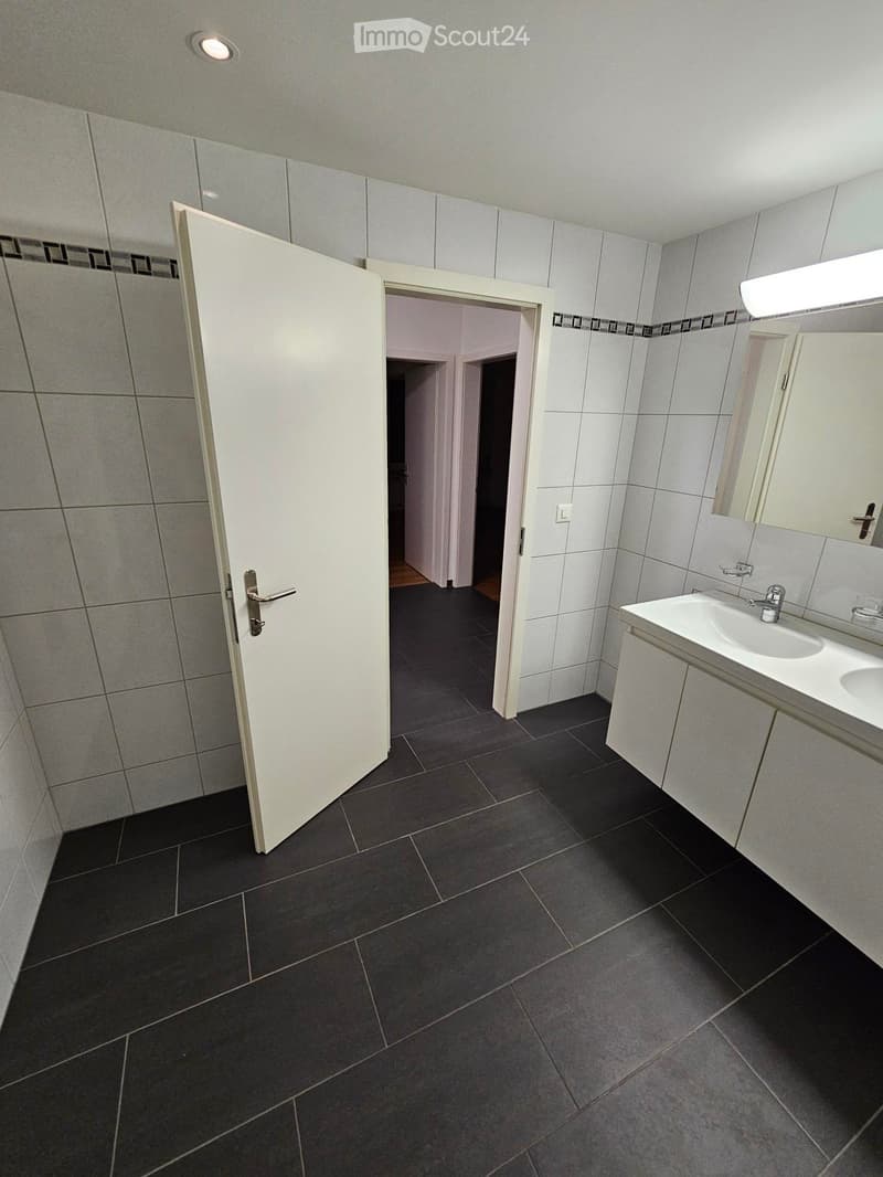 Modern bathroom with dark tiled floor, white tiled walls, a sink with a mirror, and a cabinet. An open door leads to another room.
