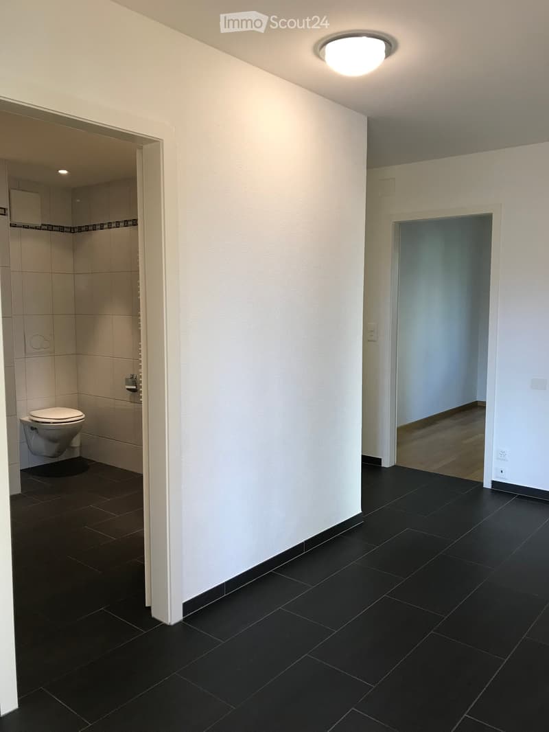 A clean, modern hallway with black tiled flooring and white walls. An open doorway leads to a bathroom with a toilet and tiled walls.