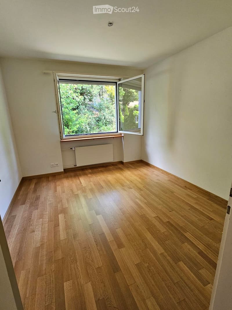 Empty room with light wooden flooring, white walls, and a large window with open shutters showing greenery outside.