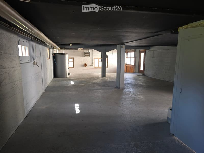Empty concrete corridor with exposed pipes, columns, and small windows allowing natural light. Features a water tank and a closed door at the end.