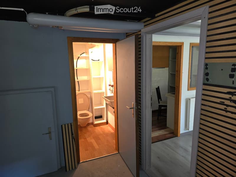 Empty hallway with open doors leading to a small bathroom and a room with a sink and chair. The bathroom has a toilet, sink, and shelves, while the adjacent room contains a sink and a chair.
