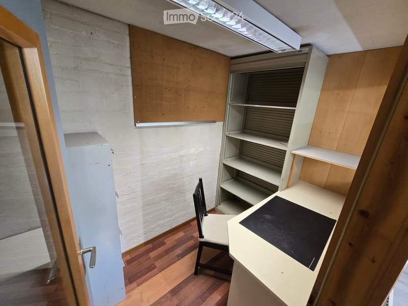 A small, furnished room with wooden flooring, a white brick wall, a desk with a black surface, a chair, and built-in shelves.
