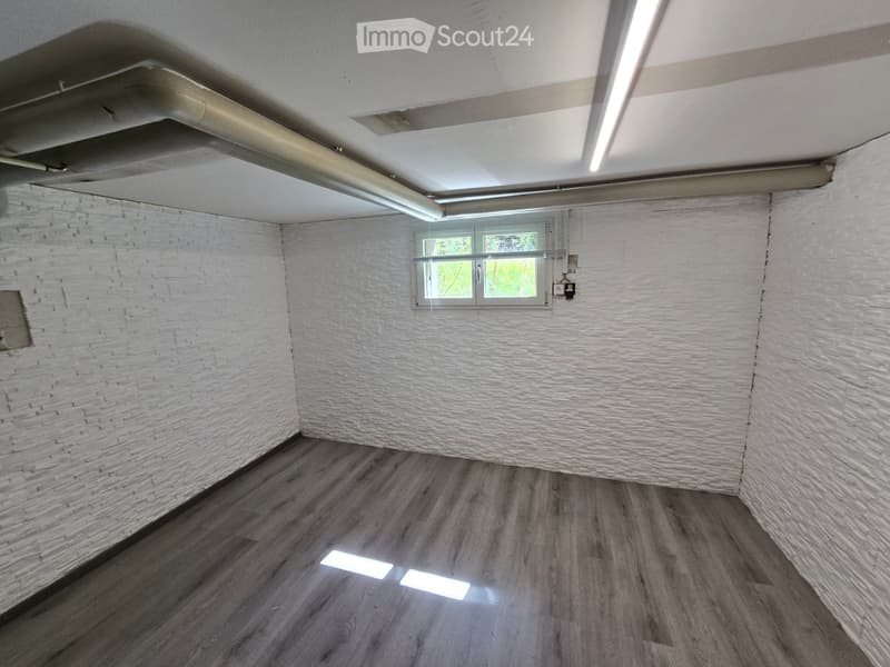Empty room with white brick walls, wooden flooring, and a single window with a metal frame. Exposed ceiling pipes and lighting fixtures are visible.
