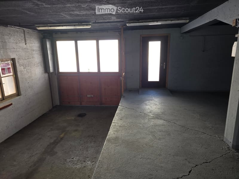 Empty concrete-walled room with a large garage door, a smaller door, and windows allowing natural light. The floor shows visible cracks and stains.