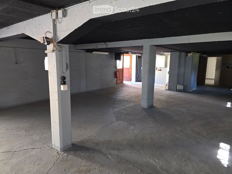 Empty underground parking area with concrete floor, white support pillars, and minimal lighting. Features a fire extinguisher and a few electrical boxes on the walls.