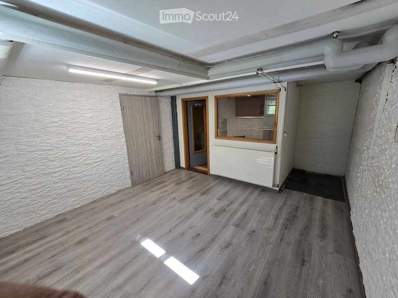 Empty room with wooden flooring, white brick walls, and exposed ceiling pipes. Features a wooden door, a small windowed area, and a radiator.