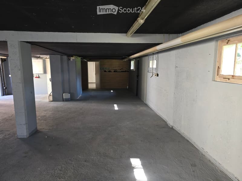 Empty underground garage with concrete floor, white walls, and minimal lighting. Features include a few pillars, a small window, and a closed garage door in the background.