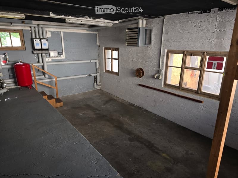 Empty basement or utility room with concrete floor, grey walls, exposed pipes, a red water heater, and small windows.
