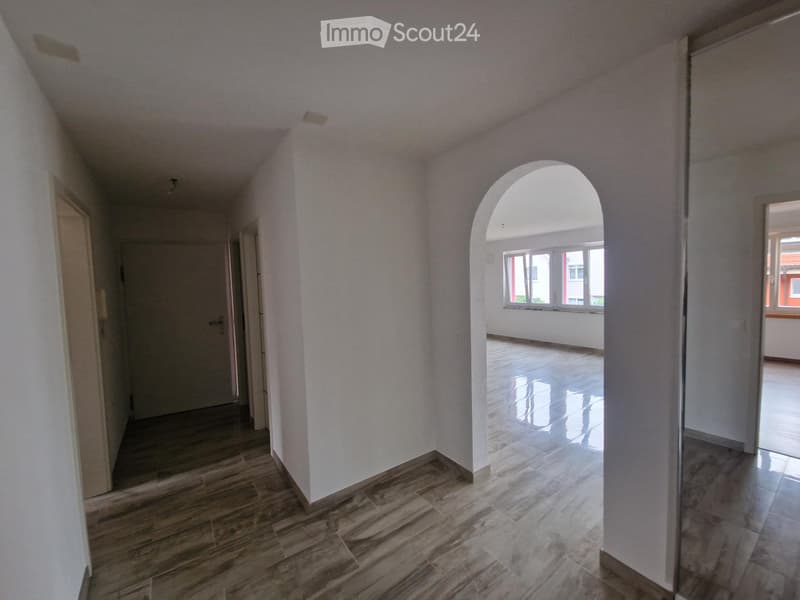 Empty hallway with light-colored walls, wooden flooring, and an arched opening leading to another room with large windows.