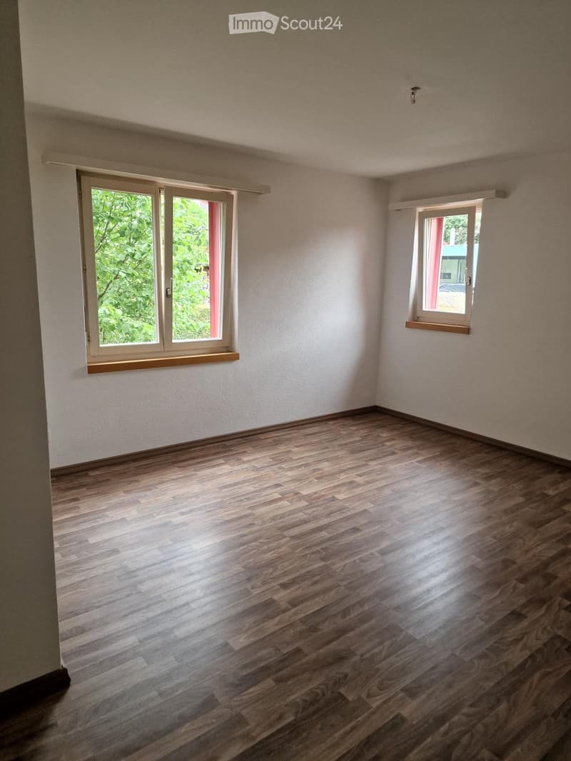 Empty room with wooden flooring and two windows, one with a view of greenery outside.
