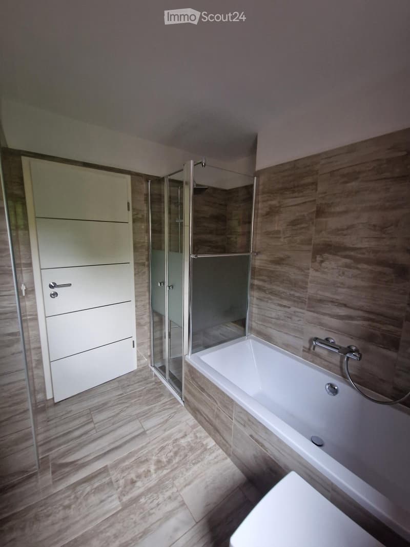 Modern bathroom with a bathtub, toilet, and a glass-enclosed shower. The walls are tiled in a wood-like pattern, and the floor is made of light-colored tiles.