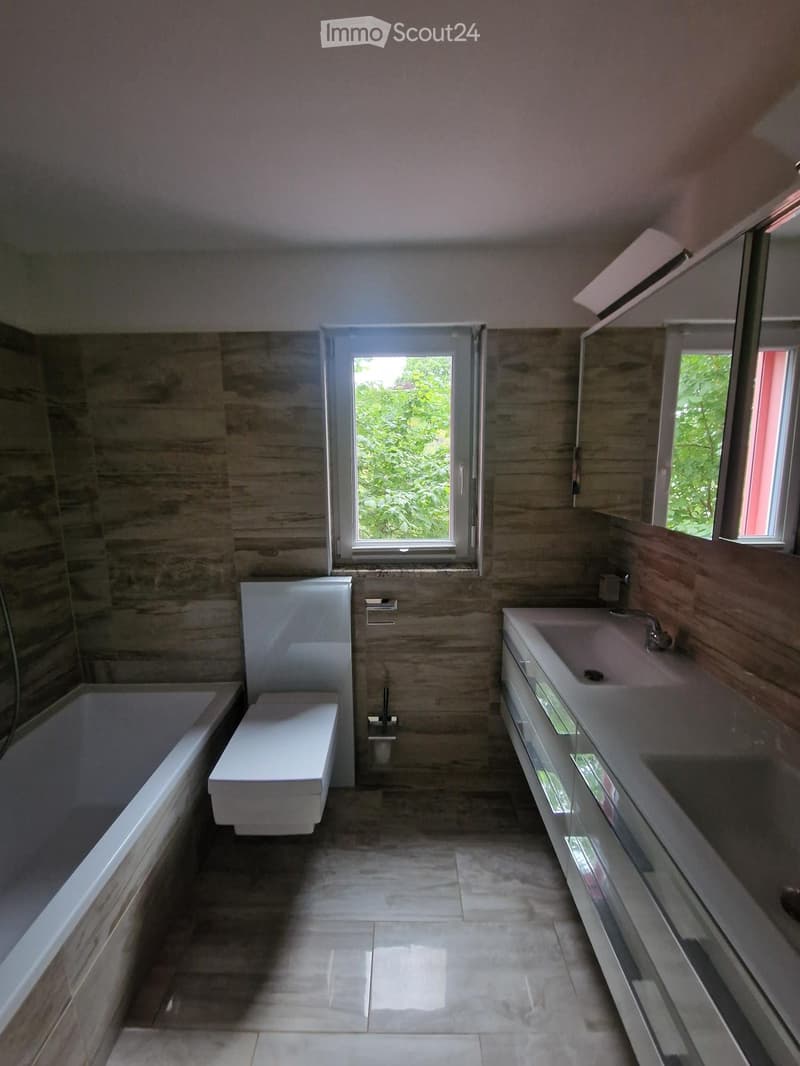 Modern bathroom with a bathtub, toilet, sink, and vanity cabinet. Features large windows with a view of greenery outside.