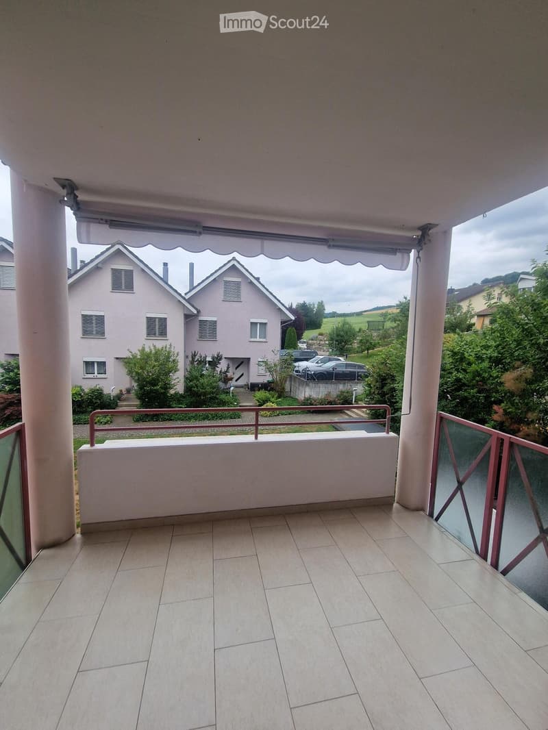 Balcony with tiled floor, white walls, and a covered awning. The view shows a residential area with houses, parked cars, and greenery.